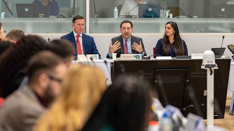 Panelist speaking and gesturing at a table alongside two colleagues during a public meeting.