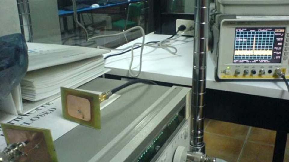 A lab workbench with a patch antenna connected via coaxial cable to a spectrum analyzer, alongside a vertical metallic rod antenna.