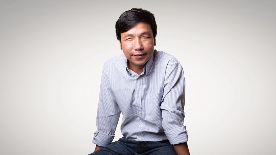 Portrait of Dang Hoai Phúc seated on a wooden block against a neutral background, wearing a light striped shirt and dark jeans, facing the camera
