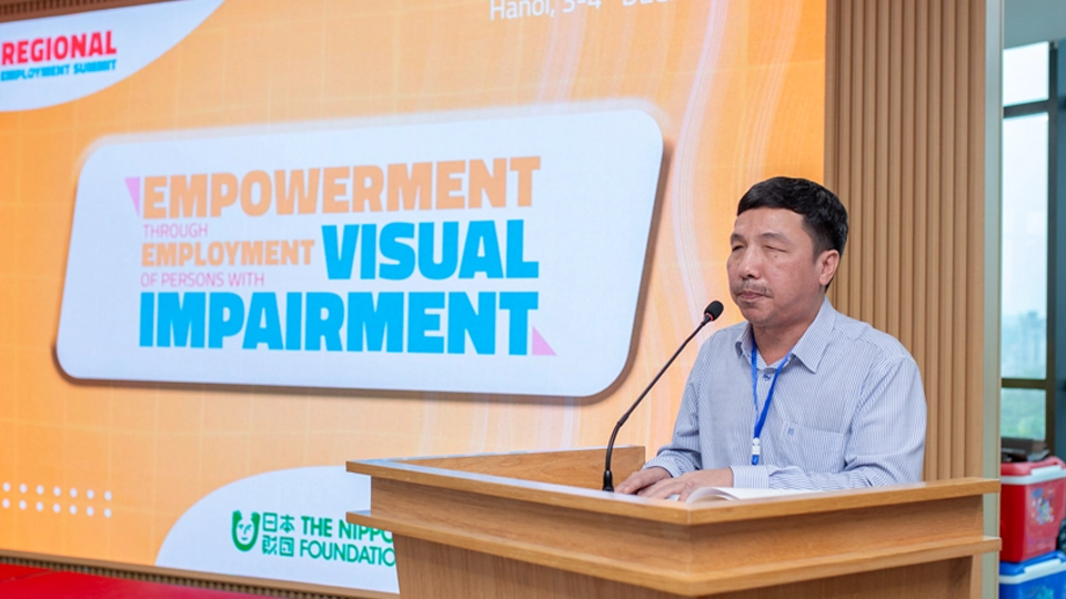Dang Hoai Phúc standing at a podium with a microphone, speaking in front of a large screen displaying “Empowerment through Employment of Persons with Visual Impairment” at a conference in Hanoi