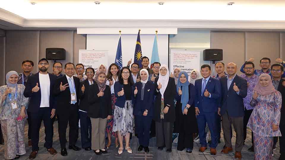 Large group of training participants standing together and giving thumbs-up in front of the Malaysian flag and event banners