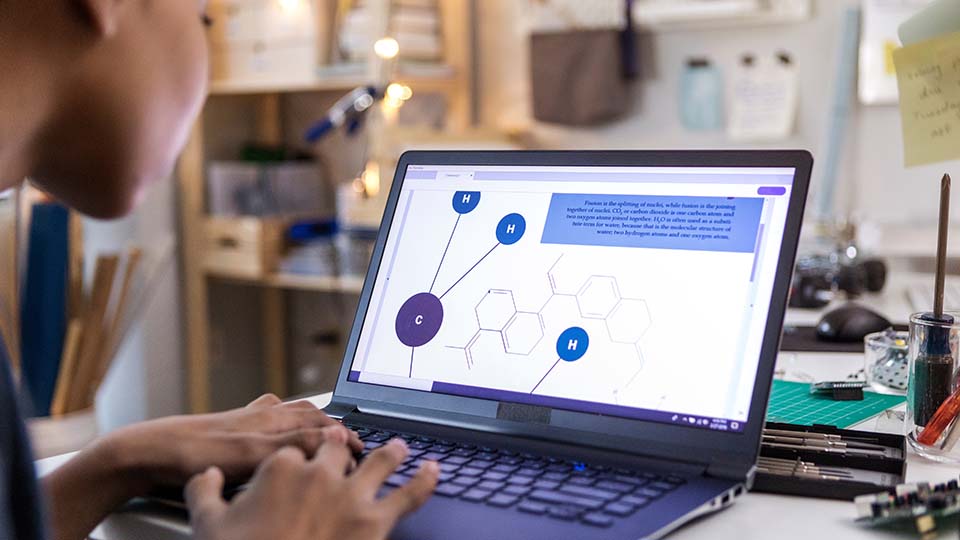 Person typing on a laptop displaying a molecular structure diagram with labeled carbon (C) and hydrogen (H) atoms, alongside text about fission and fusion.