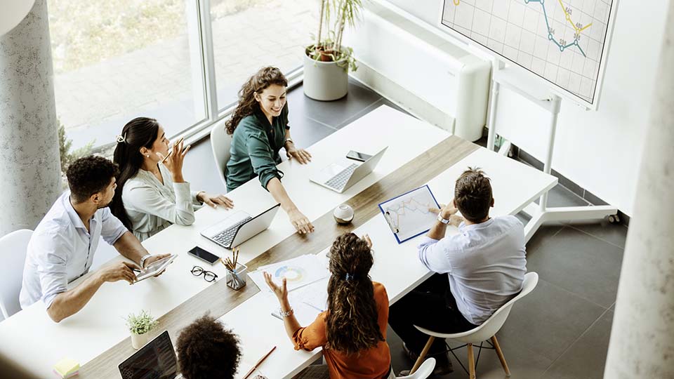 Six professionals are gathered around a white table, engaged in discussion with laptops, tablets, and printed charts spread before them.