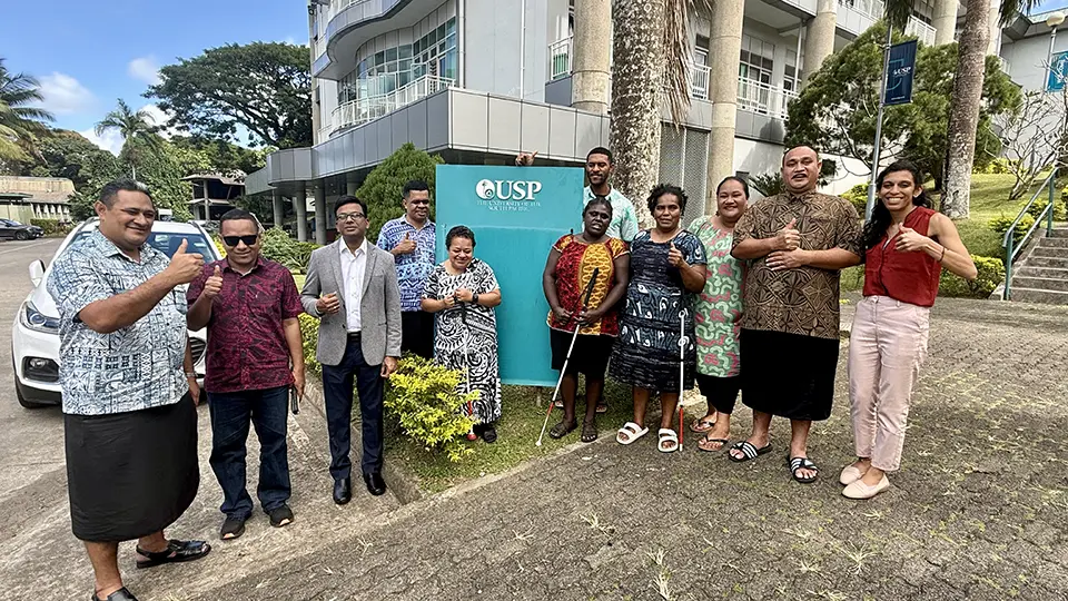 Experts and participants of the accessible book production training stand smiling and giving thumbs up in front of a sign for the University of the South Pacific (USP).