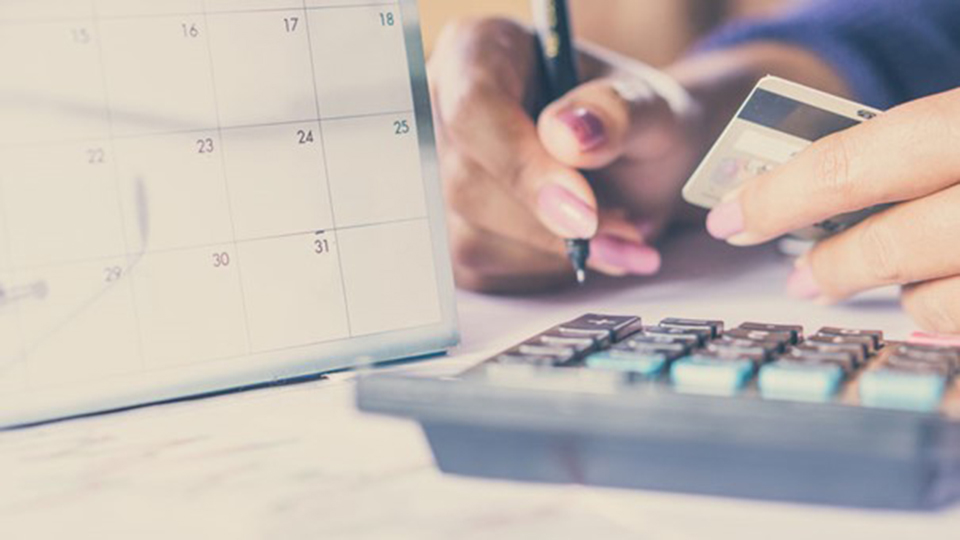 Person holding a credit card and pen while using a calculator near a calendar, suggesting financial planning or bill payment.