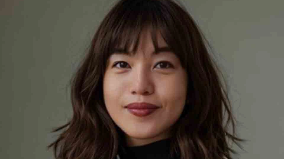 Portrait of a woman with shoulder-length wavy brown hair and bangs, wearing a black top, smiling softly against a neutral gray background