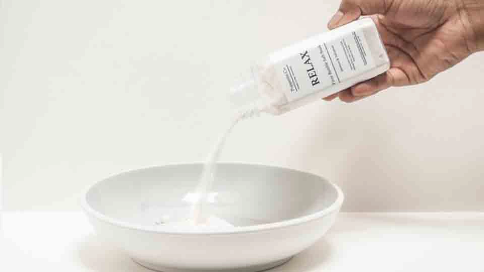 Hand pouring liquid bath salts from a bottle labeled 'Relax' into a white ceramic bowl on a white background