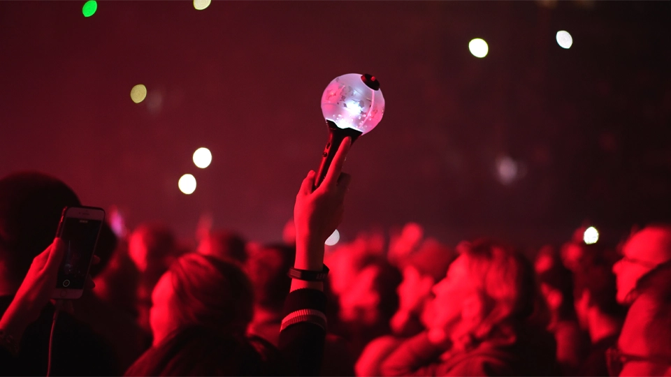 k-pop-lightstick-73531.webp Uma pessoa segura um lightstick de K‑pop no meio do público de um show, cercada por fãs sob a iluminação vermelha do palco; no fundo, há pontos de luz espalhados.