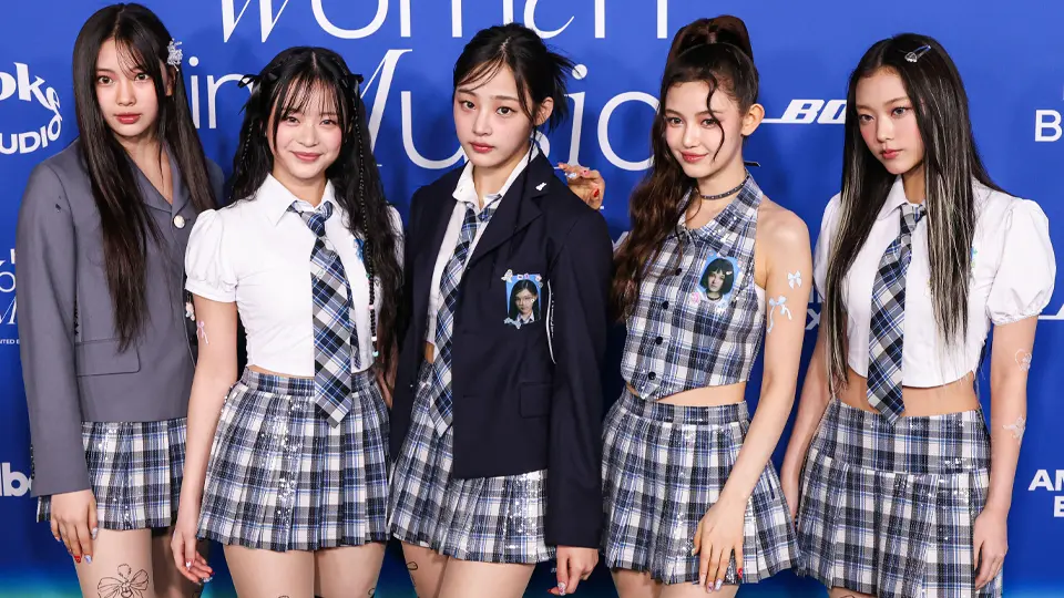 k-pop-newjeans1-73531.webp As cinco integrantes do NewJeans posam sobre um tapete azul usando roupas xadrez em estilo colegial combinando; dois integrantes estão sorrindo e as outras têm no rosto uma expressão neutra ou reservada.