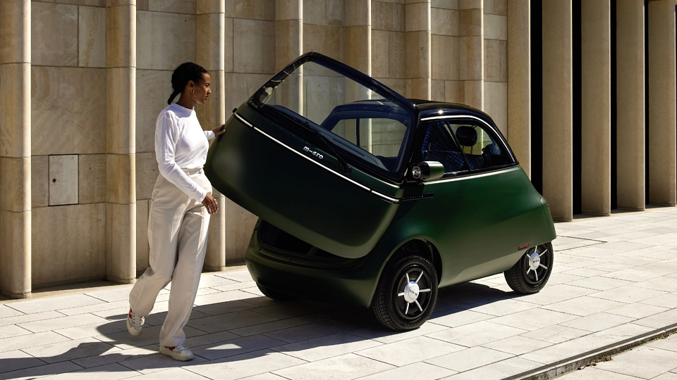 A person in light-colored clothing opens the front door of a compact dark green Microlino electric microcar, parked on a paved street beside a modern stone building with tall vertical columns