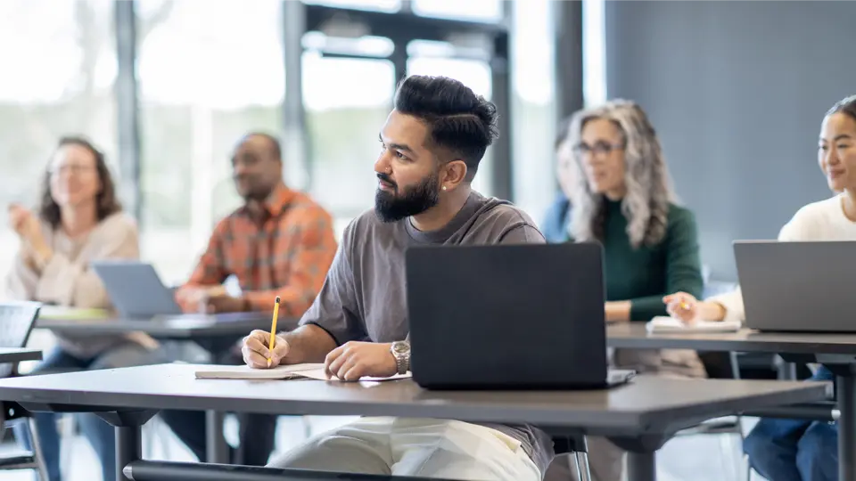 En un aula moderna y luminosa, un grupo de adultos se sienta en pupitres con ordenadores portátiles. En primer plano, una persona toma notas mientras escucha atentamente.