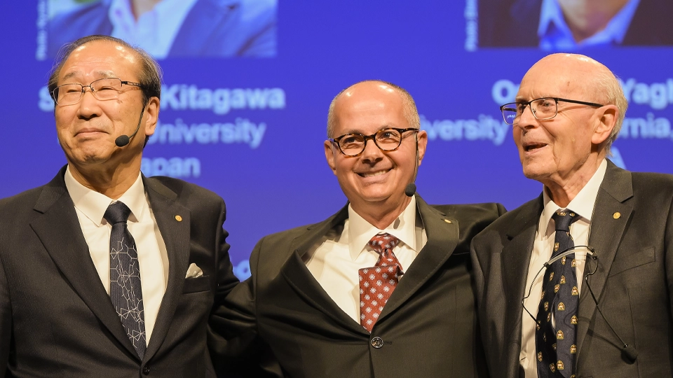 Susumu Kitagawa, Omar M. Yaghi and Richard Robson standing close together, smiling and holding each other in a friendly pose in front of an auditorium backdrop.