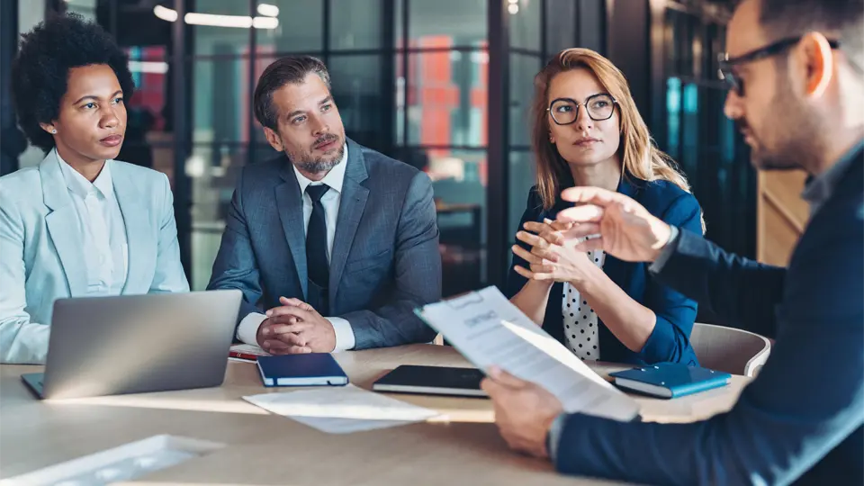 Four business professionals sit at a table in a glass-walled office during a negotiation as one person gestures with a document, and the others listen.