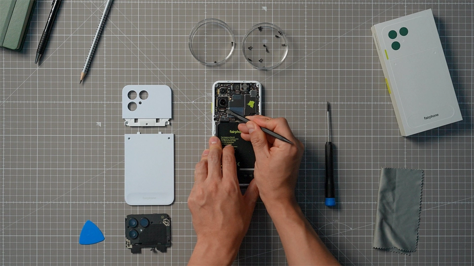 Hands use a tool on an open Fairphone with modular components laid out on a cutting mat nearby.