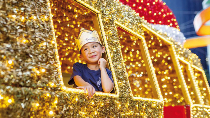 照片中，一个头戴王冠的小男孩站在一辆布满灯和亮片的马车上。