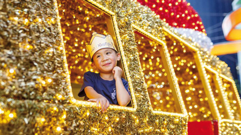 Foto de un niño que lleva una corona sentado en una carroza cubierto de luces y espumillón.