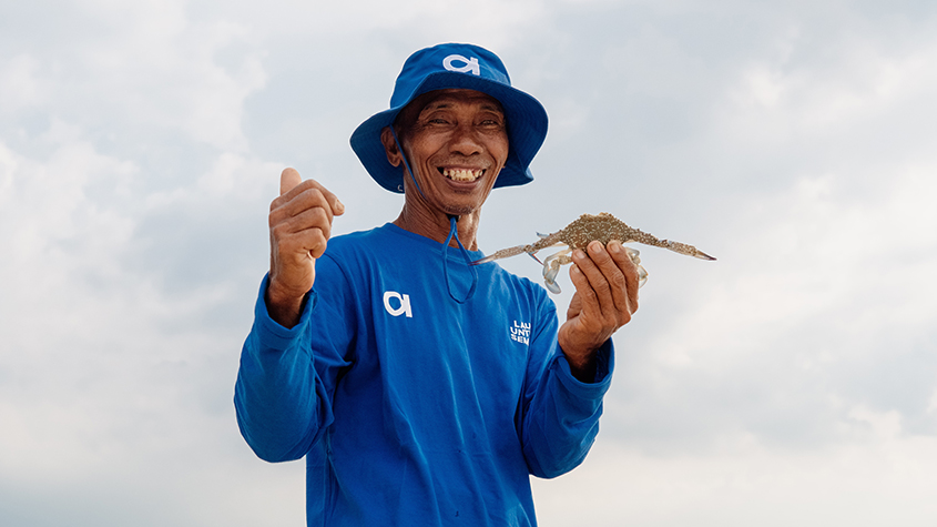 A smiling fisherman in a blue Aruna tee-shirt is holding a blue swimming crab