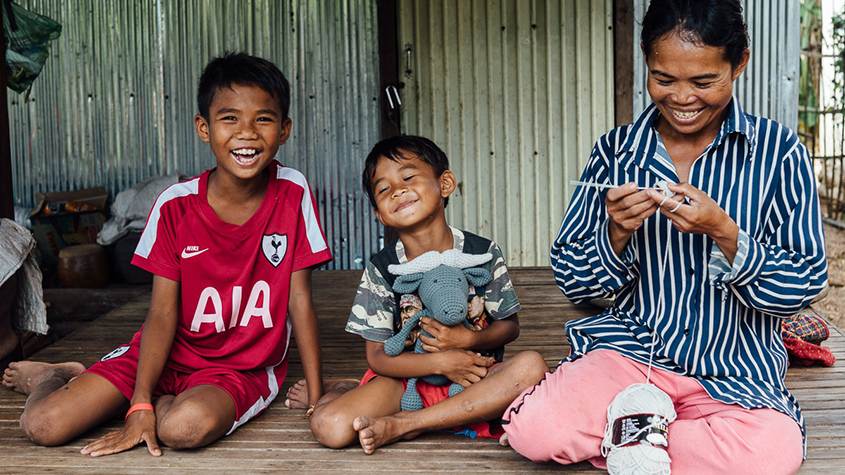 Pkorn Malis in a striped shirt sitting on the wooden floor outside and using a crochet to knit with two young children, one of them holding a Bongo, the grey knitted water buffalo