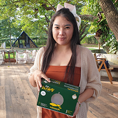 Chutima Sonloy, a “Prachin Durian” producer is standing on a wooden deck, in a garden, and holding an empty box of her products, with the yellow Thai GI logo on it