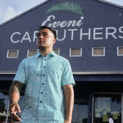 A young man wearing a light blue Elei shirt with traditional Samoan print, standing in front of a building bearing the name of Eveni Carruthers, a Samoan apparel company