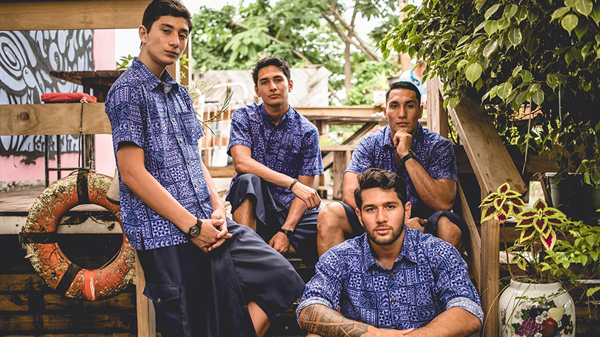 Four young men in a bucolic environment, wearing the same outfit: medium blue Elei shirts and dark blue Ie Faitaga (traditional dress trousers). Three of them are sitting on metal stair steps