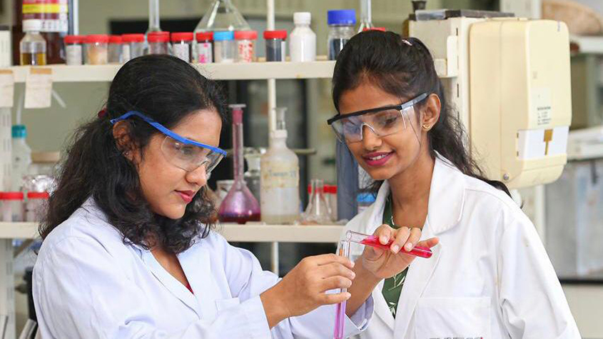 Haily Seneviratne with Danushika Demini, a research scientist, both wearing security glasses in a laboratory while Haily is transferring a pink substance from one test tube to another