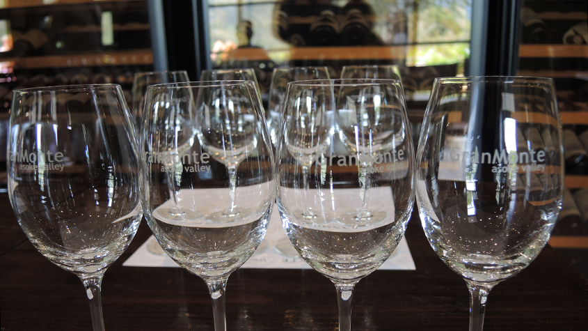 A row of GranMonte engraved wine glasses on a GranMonte place mat with in the background on each side two tall wine cellars.
