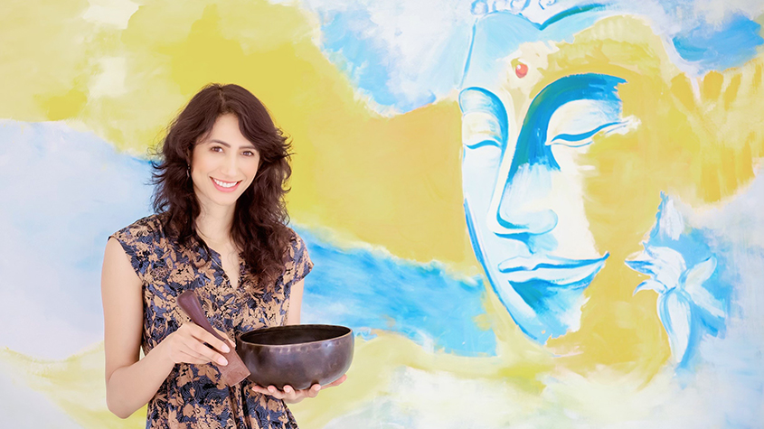 Prerana Shah, founder of Luxury Yogi, holding a chanting bowl, with a blue and yellow background featuring a peaceful face