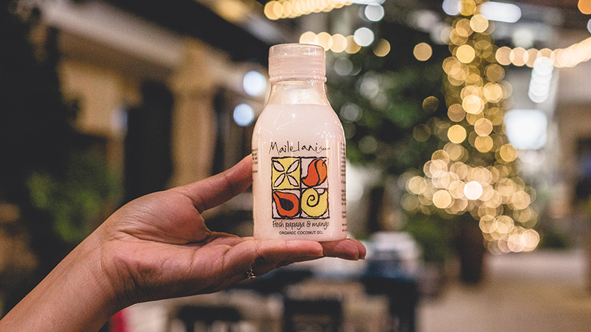 A hand holding a bottle of Mailelani coconut body oil with a colorful label mentioning fresh papaya and mango