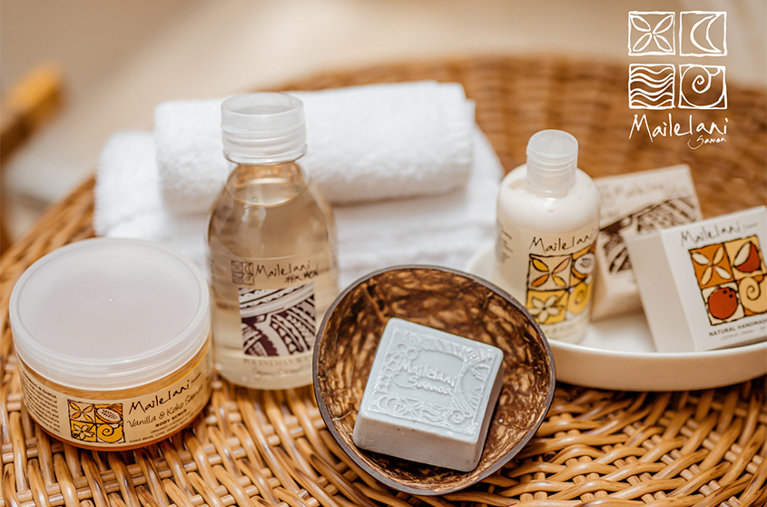 An assortment of Mailelani products: scented coconut body oil, body scrub, and soap in a woven basket