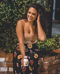 Tailani Salanoa-Chung, General Manager of Mailelani in a flowered skirt, sitting on a low brick wall and holding a bottle of Mailelani scented coconut body oil