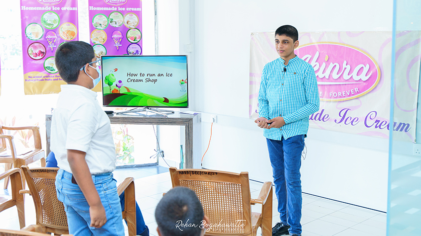 Nethila standing next to a computer screen explaining how to run an ice cream shop to a standing teenager