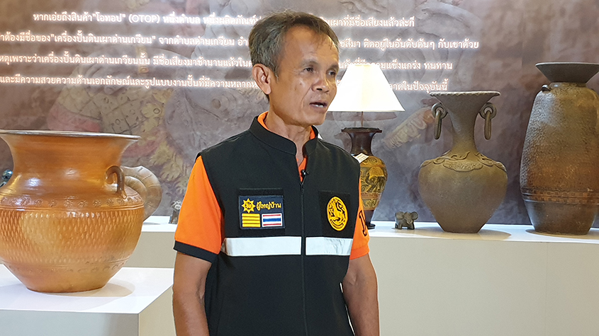 Mien Sintaleq, a potter and the leader of Dan Chai village, standing in a display room, in front of several Dan Kwian Pottery items