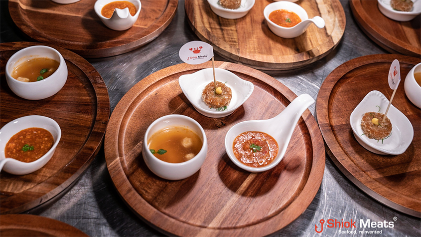 Several wooden plates with small ceramic cups containing sample cultured crustacean-based food, including crab cakes and chili