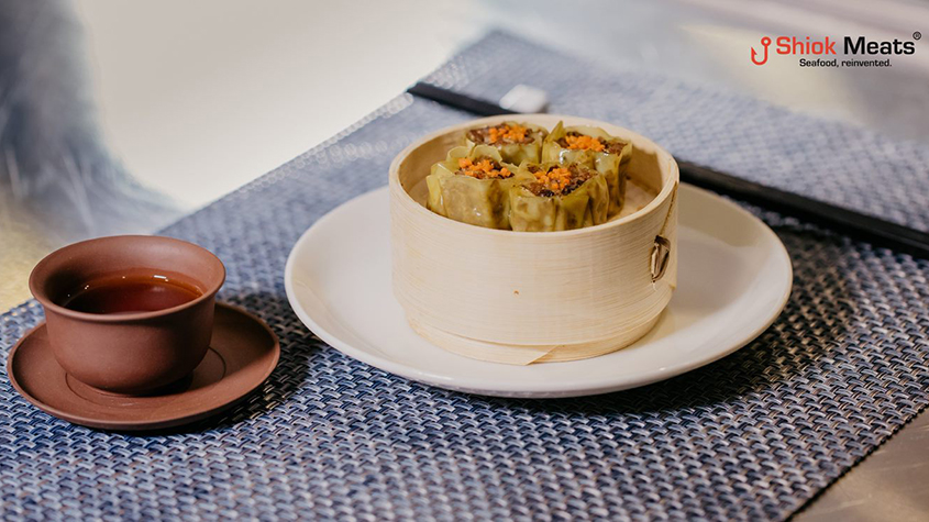 A wooden steam box containing dumplings filled with cultured shrimp meat on a mat, next to a ceramic cup of tea