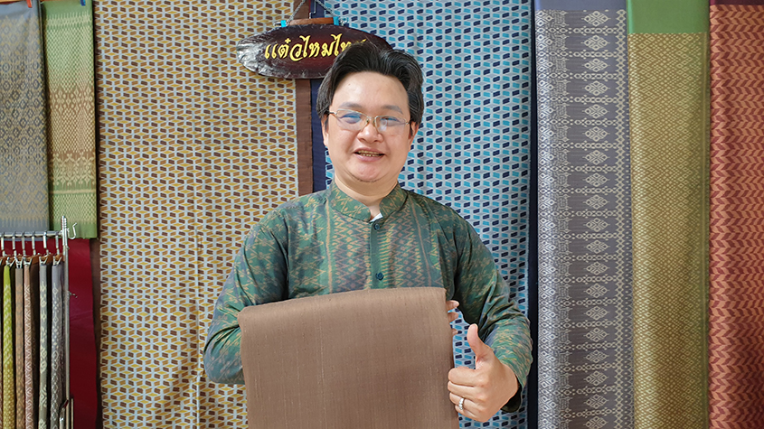 Sakchai Boonyanusith, a silk weaver and owner of Taew Thai Silk holding a piece of light brown silk, smiling with a thumb up. Several pieces of colorful and patterned silk is hanging in the background