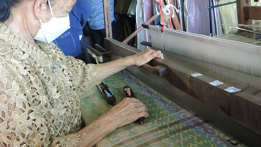 A woman working a traditional two heddles loom, weaving a green and blue pattern with two spools