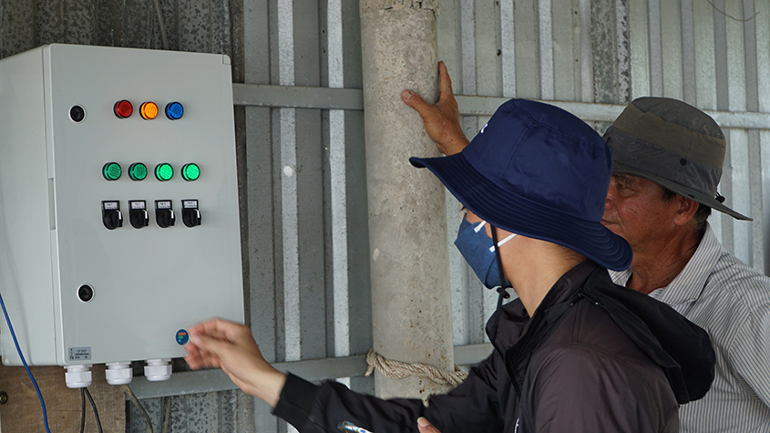 Two men with sun hats on looking at a metal electric control cabinet with several colored lighted buttons