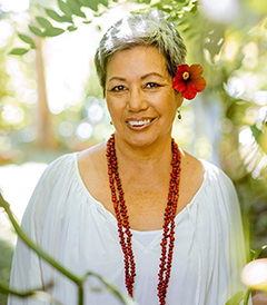 Shelley Burich, owner of Vaoala Vanilla, in a white dress, with a red flower in her hair and a long red necklace behind a vanilla plant in a garden