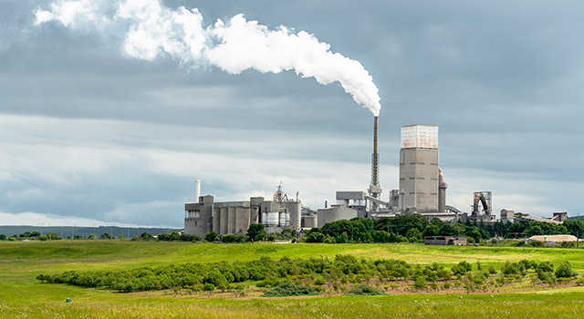 Cement factory with white smoke coming from a high chimney in the Scottish countryside on a cloudy summer day. A grassy field with sheep grazieng is visible in foreground. Dunbar, Scotland, UK: