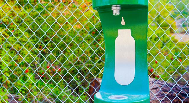 Green water bottle refill station set against a mesh wire fence outdoors. Convenient access to clean drinking water while enjoying nature.