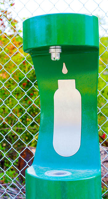 Green water bottle refill station set against a mesh wire fence outdoors. Convenient access to clean drinking water while enjoying nature.