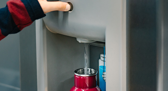 Close up of hand with long sleeve red blue stripe shirt pressing button of drinking water filling station at the Airport to refill red insulated reusable water bottle.