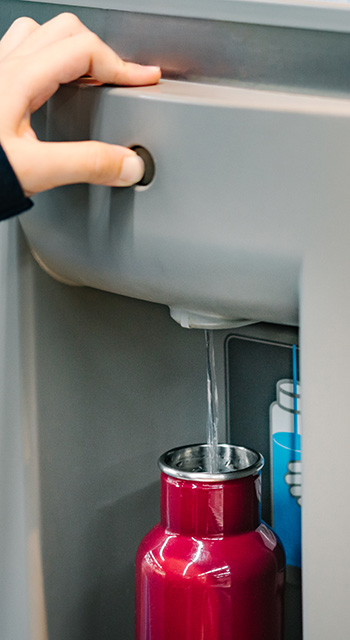 Close up of hand with long sleeve red blue stripe shirt pressing button of drinking water filling station at the Airport to refill red insulated reusable water bottle.